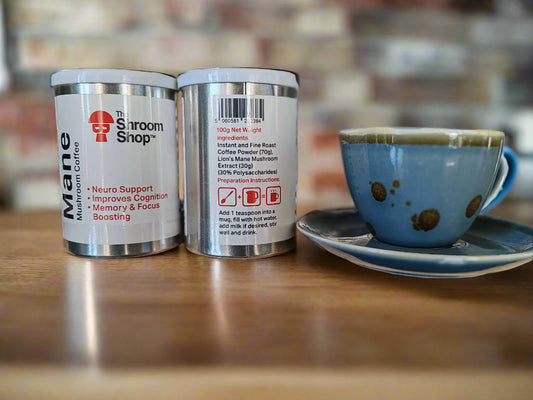 Two canisters of 'Lions Mane' and 'Shroom Shop' coffee on a wooden surface with a blue cup in the background.